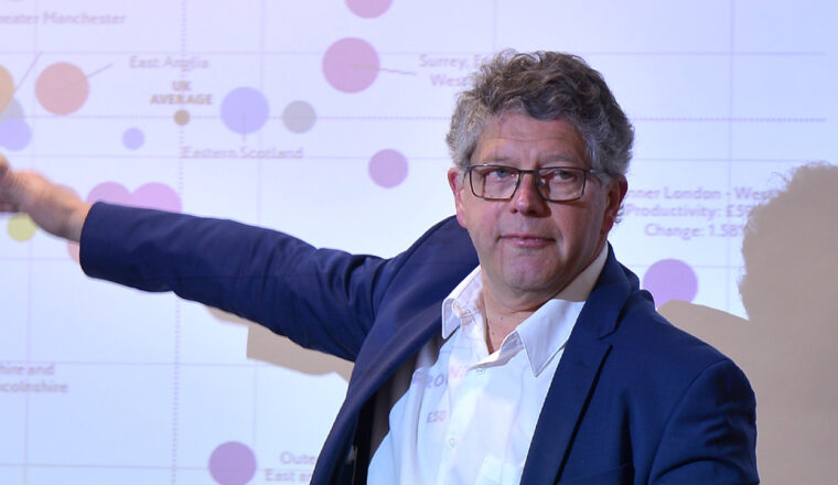 Speaker presenting during a conference session, pointing towards a slide displayed on a screen.