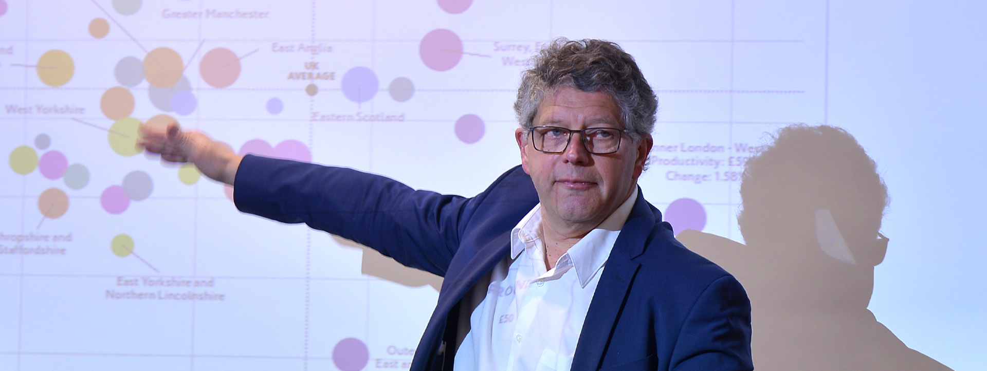 Speaker presenting during a conference session, pointing towards a slide displayed on a screen.
