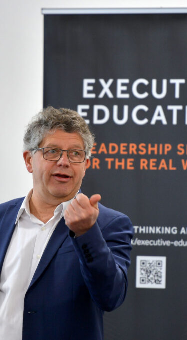 Speaker in a blue jacket gesturing during a presentation, standing in front of an executive education banner.
