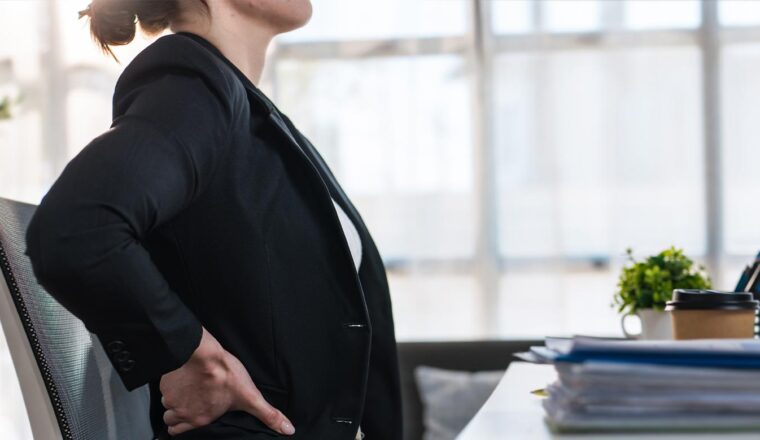 Office worker seated at a desk with one hand on their lower back, illustrating physical discomfort or pain at work.